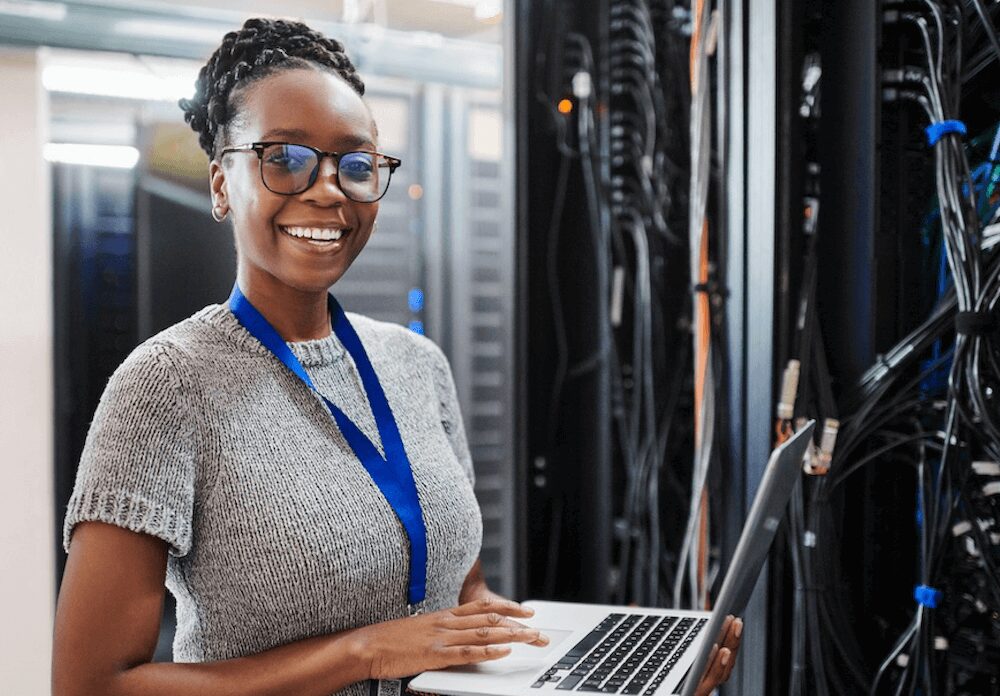 Woman holding a laptop and smiling at camera while working in a server room