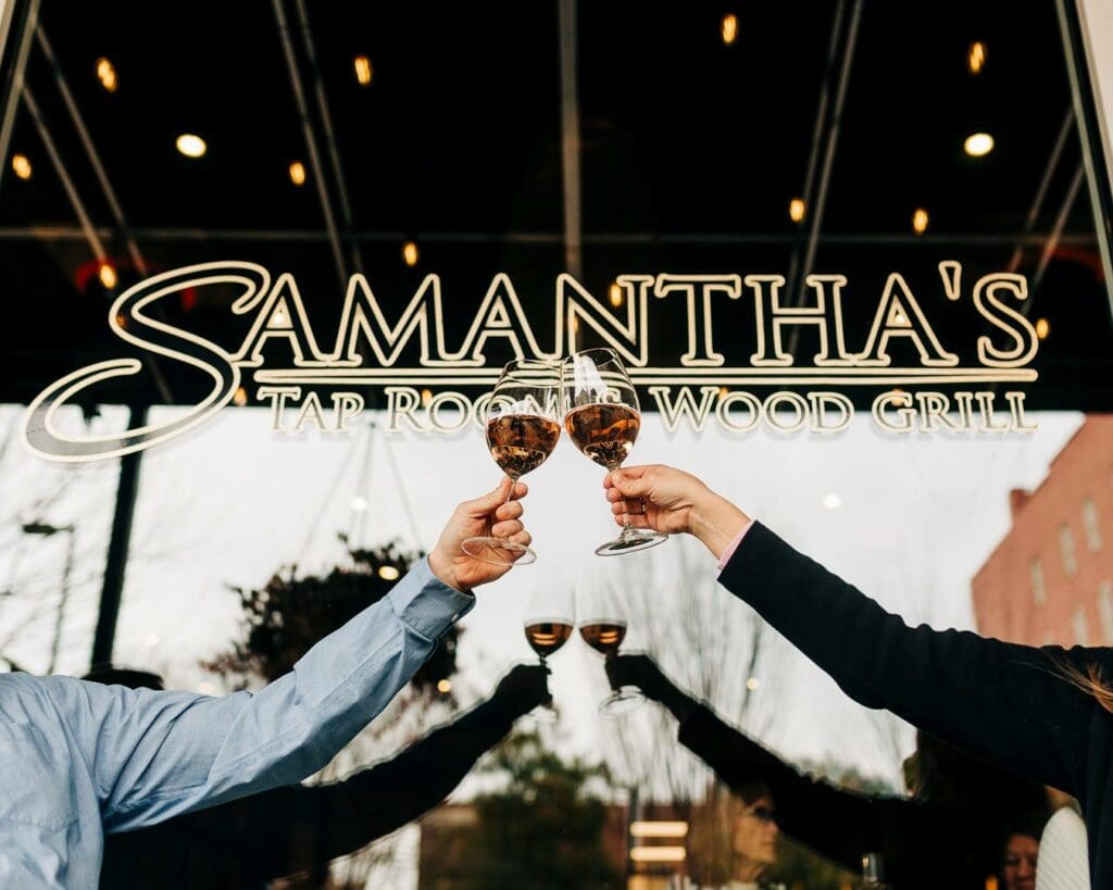 Two people clinking two wine glasses together in front of a glass window that says Samantha's